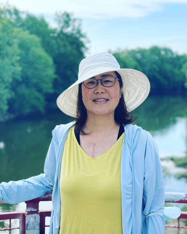 Motoko smiling outdoors by a river, wearing a sun hat